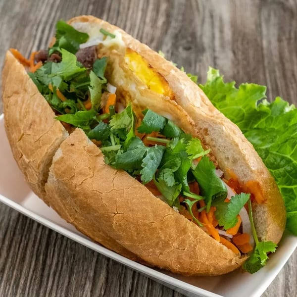 A Vietnamese banh mi sandwich filled with meat, fried egg, cilantro, and pickled carrots on a white plate.